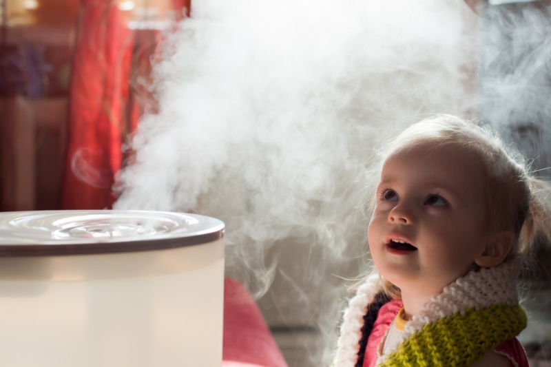 Benefits of Using a Humidifier in Your Home. Image is a photograph of a baby girl looking mystified at a humidifier.